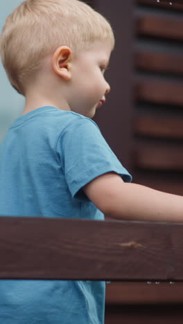 cute little boy in loose t-shirt walks touching brown wooden beam of railing along terrace deck at cottage house close view slow motion