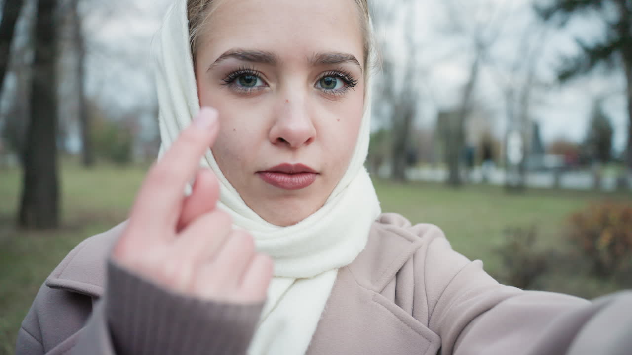 Giovane donna con la sciarpa che fa un cuore con le dita, sguardo dolce e invitante in un parco invernale, selfie ravvicinato con un cappotto accogliente, trucco delicato e sfondo bokeh, delicato movimento della mano verso la fotocamera, atmosfera da influencer spontanea.