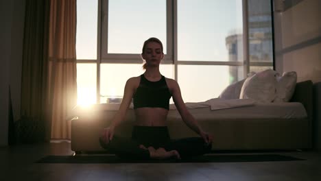 gorgeous scene of blonde female adult meditating in lotus position at home sitting on yoga mat, enjoying exercises. young people, healthy lifestyle and interiors concept. mild sunset on the background window