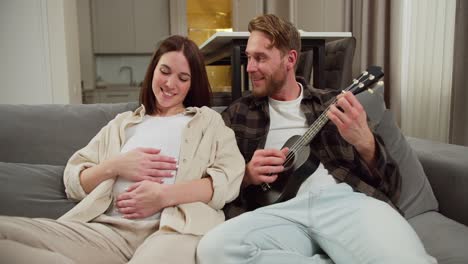 happy man with brown hair and stubble in a checkered shirt plays a black ukulele while his brunette pregnant wife sits on the sofa and listens to music while stroking her belly in a modern apartment during the day