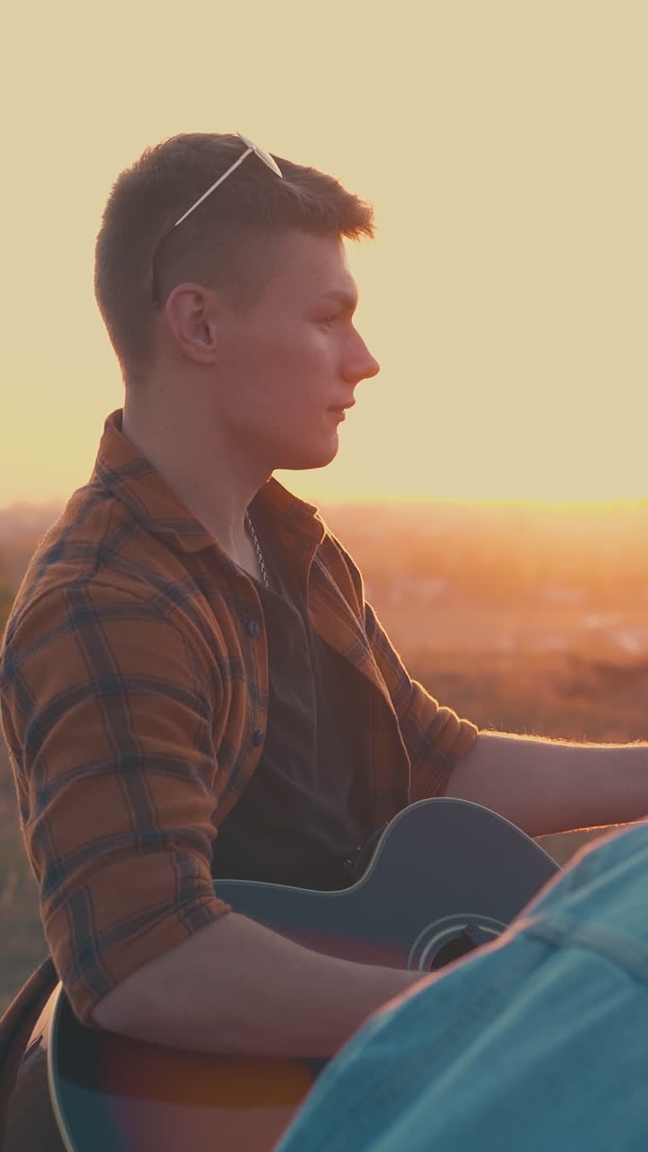 young man in glasses plays guitar to happy friends near camp bonfire in warm autumn evening against setting sun