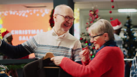 Joyful-older-couple-using-smartphone-to-chat-with-nephew-in-online-videocall-while-shopping-for-clothes,-asking-for-advice-on-picking-perfect-xmas-gift-during-Christmas-holiday-season