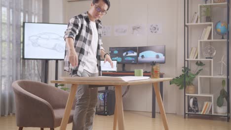 아시안 남성이 앉아서 tv와 컴퓨터 디스플레이 3d 전기 자동차 모델과 함께 스튜디오에서 자동차 디자인 스케치에 대한 논문을 작업합니다.