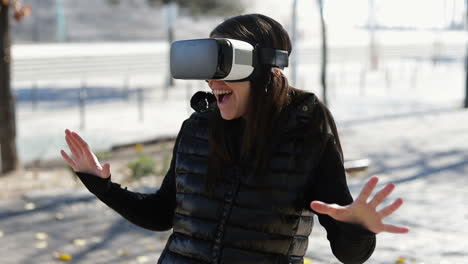 Amazed-young-dark-haired-woman-in-warm-black-waistcoat-wearing-virtual-reality-glasses-in-early-autumn-park