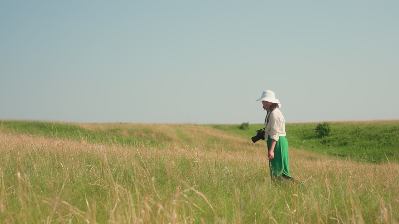 Smiling lady in white hat and green skirt holding camera walks slowly across tall golden grass in open sunny field with distant green slopes and clear blue sky creating calm scenic summer atmosphere