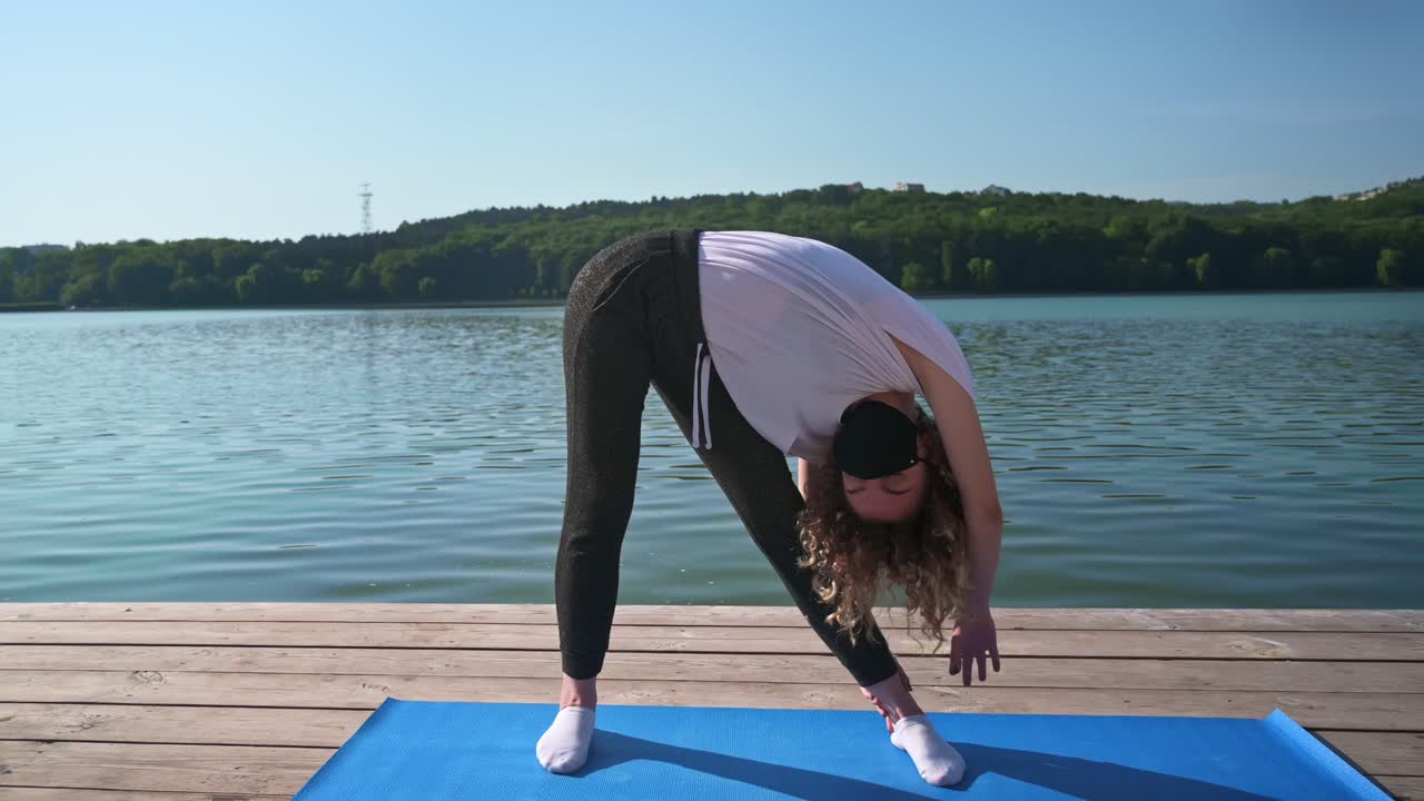 Young curly woman practicing yoga on the mat while wearing black protective mask. Park and lake in background. Sunny day. Corona Virus idea. Chisinau, Moldova