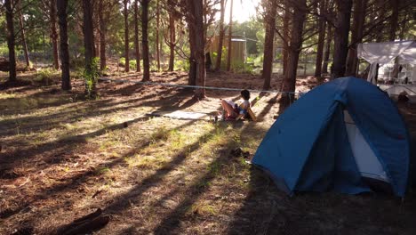 A-young-woman-is-reading-a-book-near-the-tent-at-a-campsite-in-a-large-pine-forest