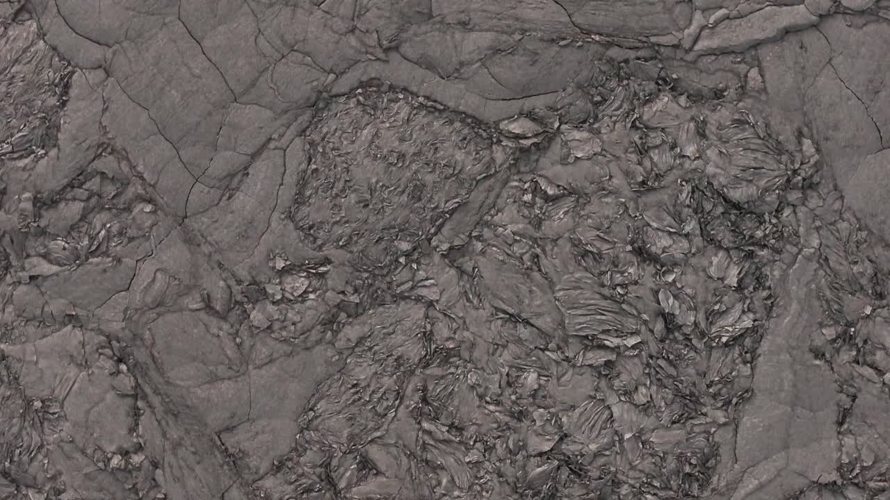 A top-down aerial view reveals the intricate textures of the Icelandic ground, showcasing a diverse landscape of rugged volcanic rock, dark earth, and remnants of snow or ice