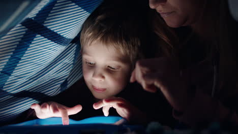 madre y hijo felices usando una tableta bajo una manta niño pequeño jugando juegos en tecnología de pantalla táctil con mamá relajándose divirtiéndose antes de acostarse