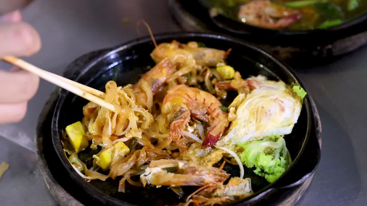 Hand uses chopsticks to mix steaming pad thai noodles, shrimp, vegetables on sizzling hot plate