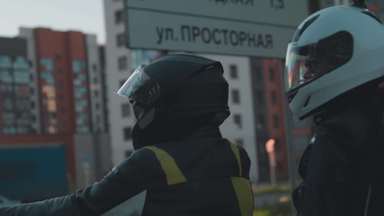 Motorcycle riders wearing helmets follow large truck on city road during sunset, warm light reflecting on asphalt and vehicles with trees, traffic signs, and buildings in background