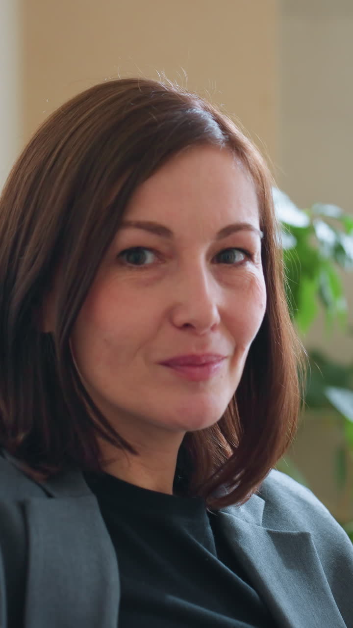 Confident businesswoman in gray blazer sitting on couch smiling warmly in modern office with indoor plants and natural lighting suggesting calm professional environment and approachable demeanor