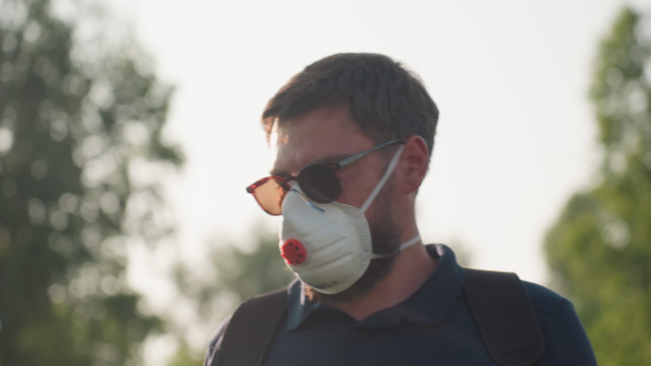 Close up gardener wearing nose mask and sunglasses looking intently at crops with soft sunlight and blurred tree background, conveying careful inspection