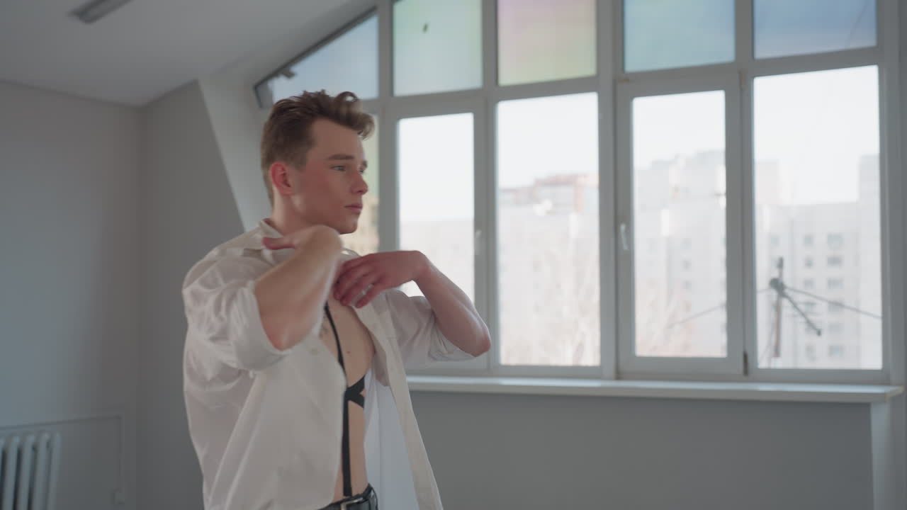shirtless boy dressed in black pants raises arms to put on white shirt in bright modern room with large window panels, natural daylight highlighting upper body against urban cityscape backdrop