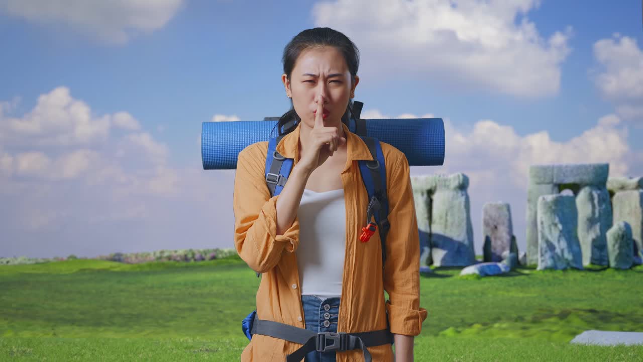 Asian Female Hiker With Mountaineering Backpack Looking At Camera And Making Shh Gesture While Traveling In Stonehenge