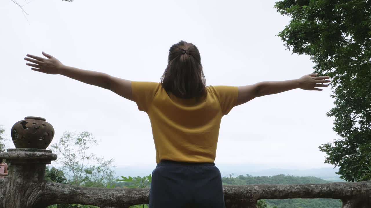 imágenes de bajo ángulo de una mujer frente al bosque y mirando hacia el cielo desde una vista alta cubierta su espalda en la cámara levantando los brazos con las palmas abiertas formando una cruz disfrutando de la naturaleza refrescante