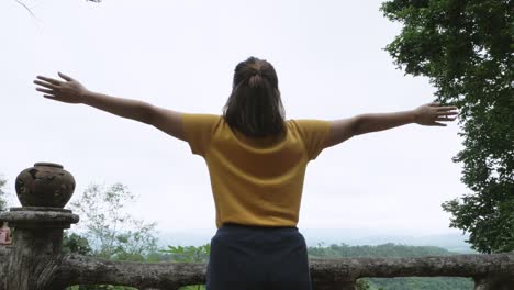 Low-angle-footage-of-a-woman-facing-the-forest-and-looking-up-the-sky-from-a-high-view-deck-her-back-on-camera-lifting-arms-with-open-palms-forming-a-cross-enjoying-the-refreshing-nature