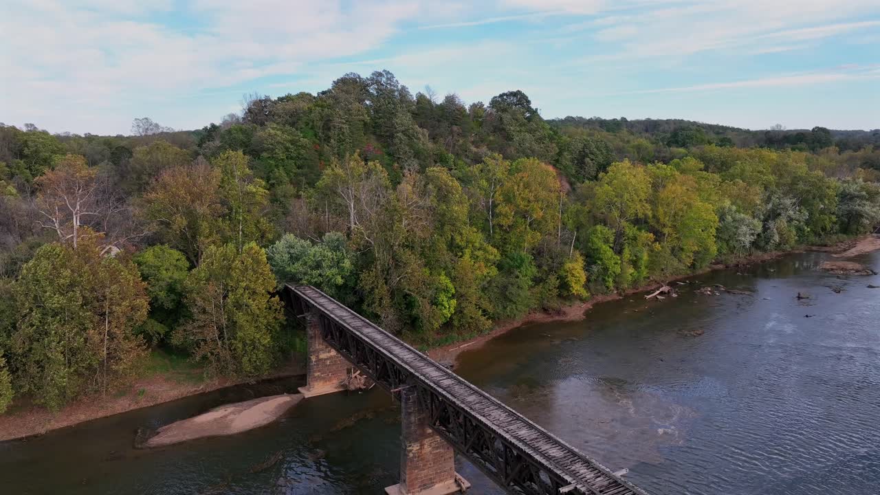 Old wooden bridge over tranquil river in forest landscape of USA. Sunset time in autumn season. Descend drone shot.
