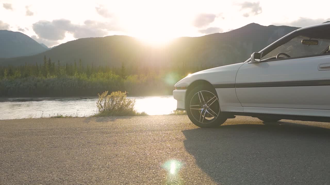 A man seen walking up to a vehicle sits on its hood to enjoy the view. An amazing lens flare is created by the setting sun.