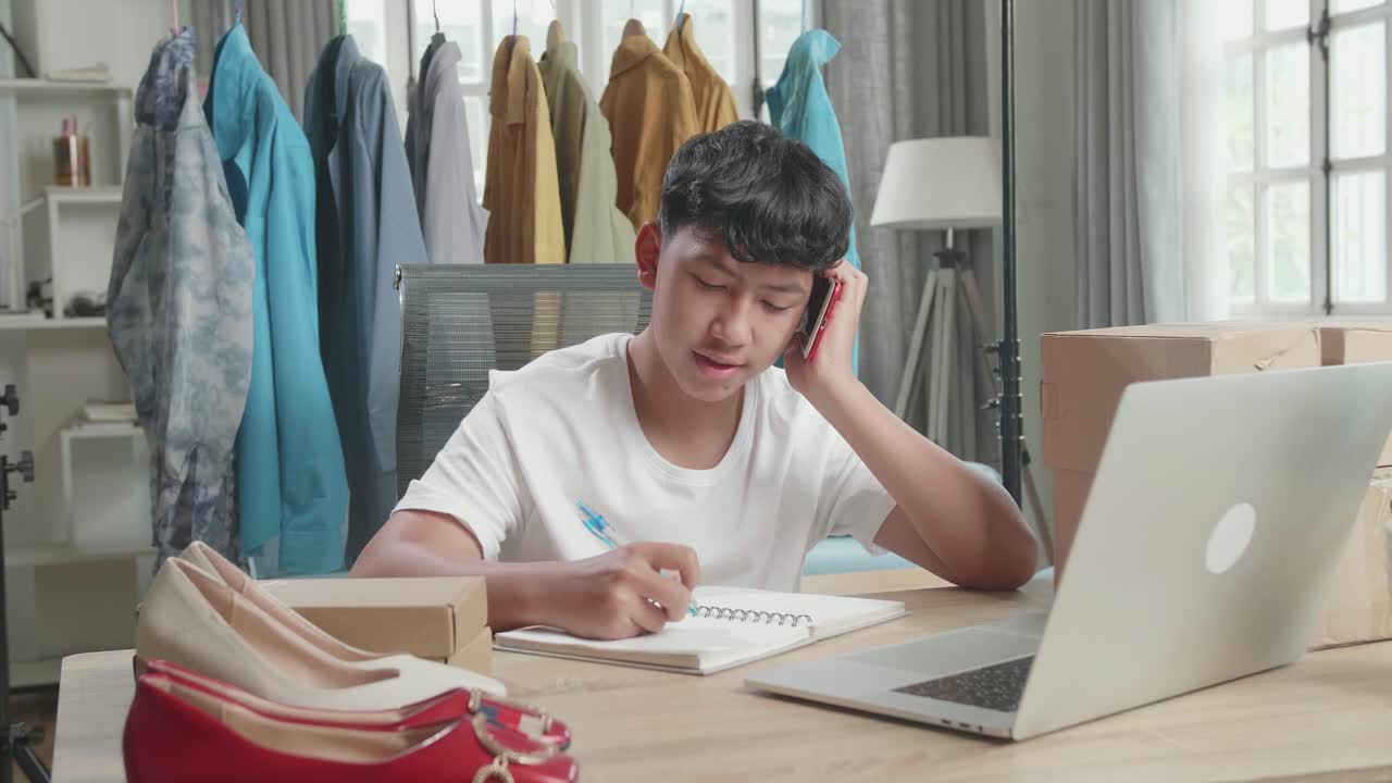 Asian Man Online Seller Talk On Mobile Phone And Take Note While Using Computer For Selling Clothes At Home