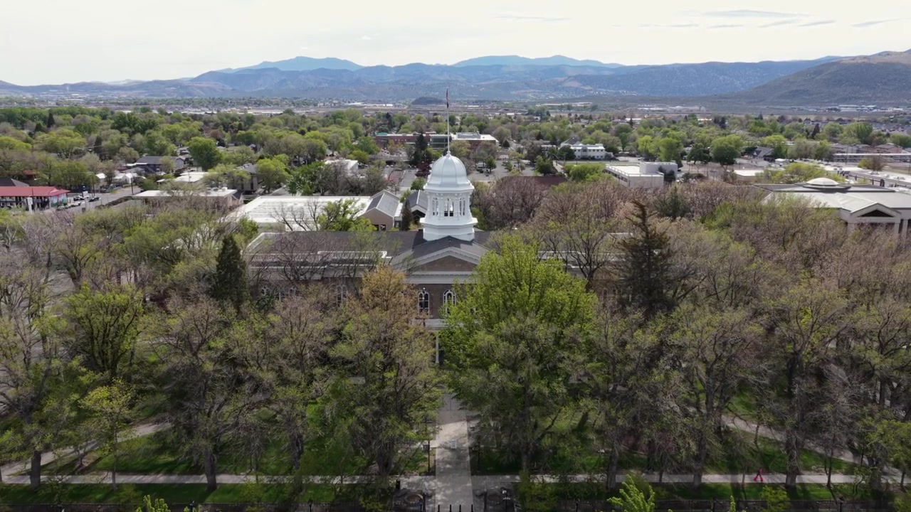 Edificio del Campidoglio dello Stato del Nevada a Carson City con cupola bianca e cupola argentata, immerso tra alberi verdi con montagne desertiche durante una soleggiata giornata primaverile negli Stati Uniti. Ampia ripresa aerea all'indietro.