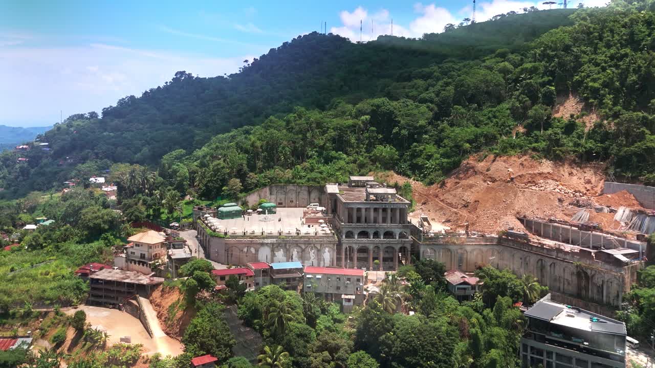 Drone footage of a large neoclassical estate under construction on a tropical hillside in the Philippines, surrounded by dense forest and scattered rural homes