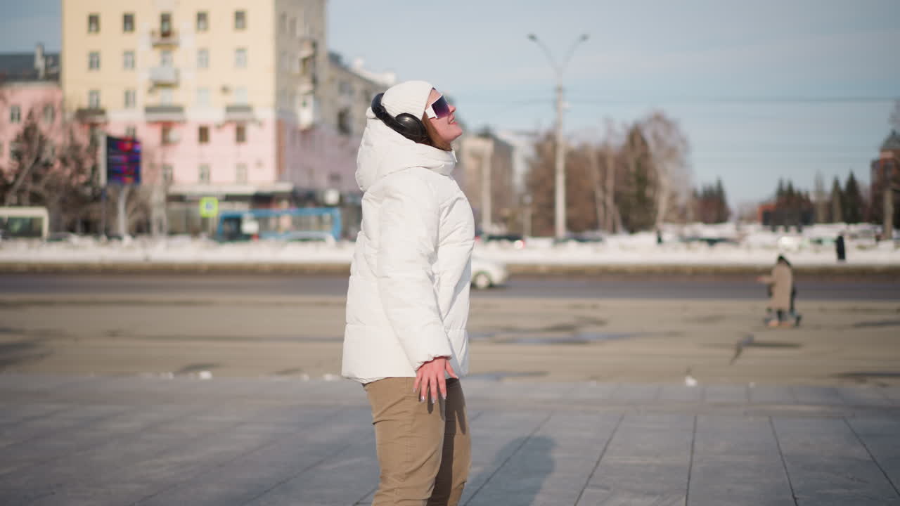 Woman wearing white winter coat, beanie and headphones passionately performing break dance moves on busy urban plaza framed by tall trees surrounded by snow, passing cars and pedestrians