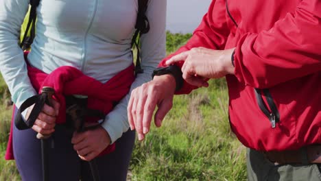 mid section of senior hiker couple with backpacks and hiking poles using smartwatch while hiking