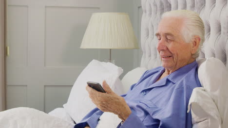 Hombre-Mayor-En-Casa-Usando-Pijamas-En-La-Cama-Haciendo-Videollamadas-En-El-Teléfono-Móvil