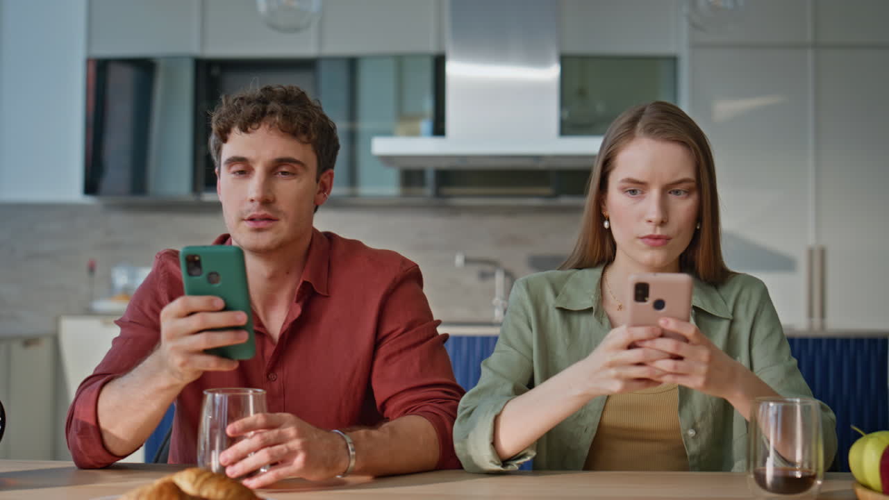 Separate couple looking smartphones sitting at apartment kitchen table closeup