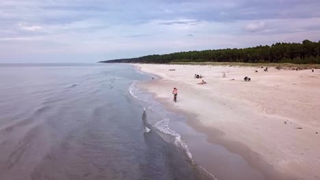 Toma-Aérea-De-Un-Hombre-Montando-En-Bicicleta-En-Una-Playa-De-Arena-En-Un-Día-Nublado