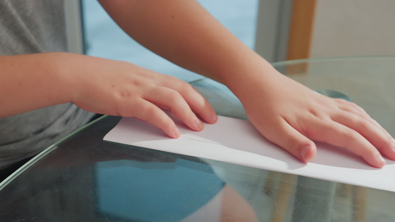 Close-up view of child hands folding and joining paper on glass table, focusing on the careful process of folding and aligning the paper for crafting project