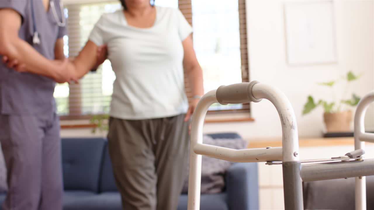 Helping senior woman walk, male physiotherapist supporting her with walker in living room