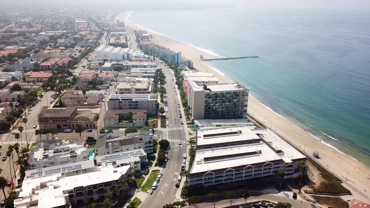 Drone pushes in along the Redondo Beach oceanfront palm lined boulevard beachfront hotels apartments and wide sandy shoreline on a late summer day. Clear water gentle surf and coastal urban density