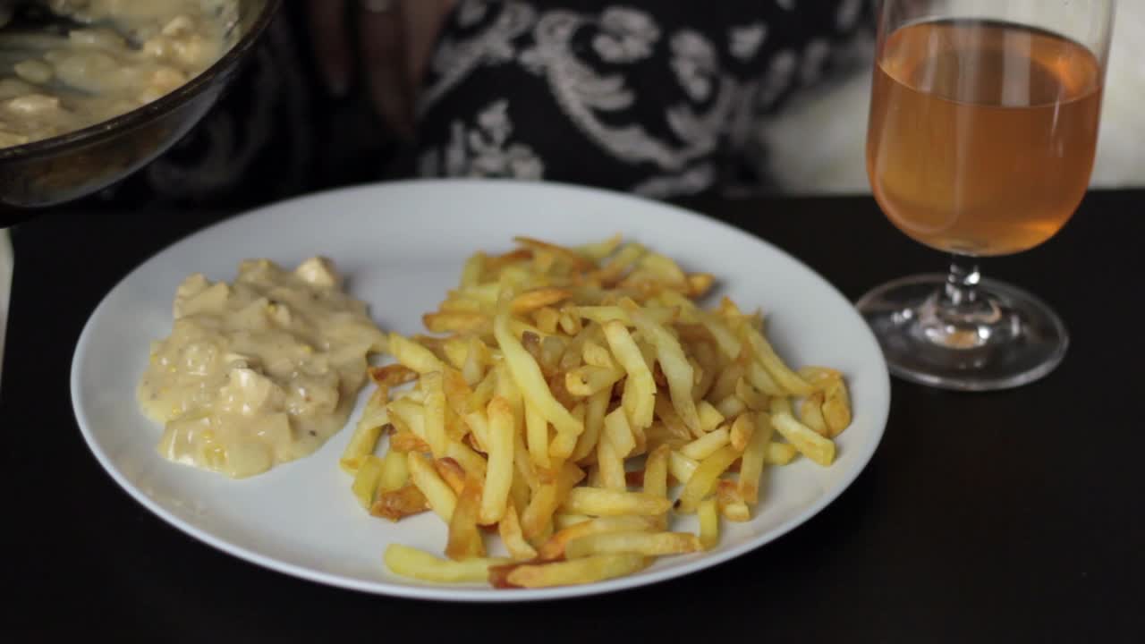 A person serving creamy sauce with a basting spoon on a plate with French fries, close up static