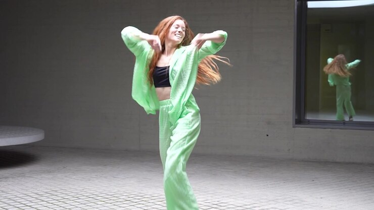 A young woman dressed in a green outfit is energetically dancing outside a modern building