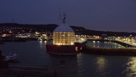 Enchanting-view-of-Howth-harbor-by-night,-close-orbit-around-lit-lighthouse,-town-lights-aglow-in-the-backdrop