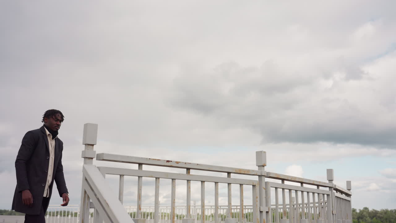 Young lad exhales while looking toward distant riverbank, standing near metal railing with calm thoughtful posture, showing peaceful emotion and quiet reflection in tranquil outdoor environment