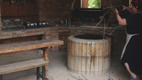 Woman-in-outdoor-brick-kitchen-adding-a-pile-of-branches-into-a-wooden-tone