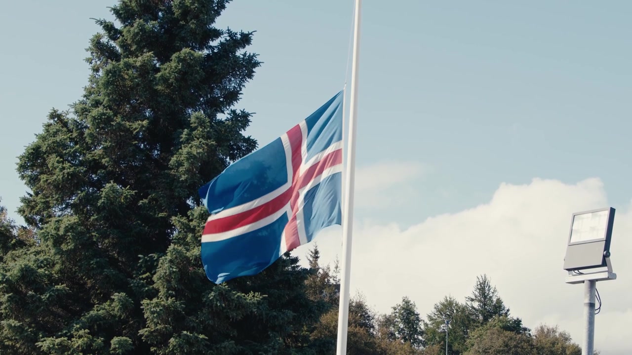 La bandiera nazionale dell'Islanda sventola al vento a mezz'asta su un'asta contro un cielo blu con alberi sullo sfondo ad Akureyri.