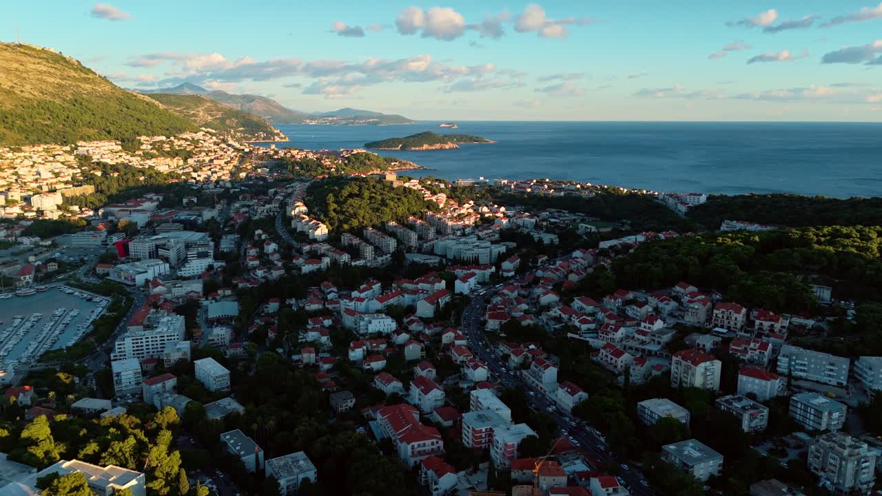 Aerial high angle drone shot flying over Dubrovnik at sunset, capturing the city’s red rooftops, surrounding mountains, and shimmering Adriatic Sea coastline glowing in soft evening light