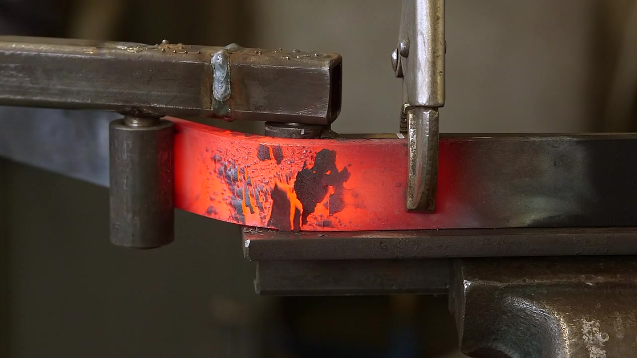 Close-up of a skilled blacksmith bending a red hot steel bar with a custom metal jig during a traditional iron forging process in a workshop.