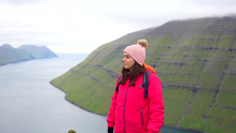 woman in pink jacket fixing her hair and beanie on top of klakkur mountain with fjord landscape