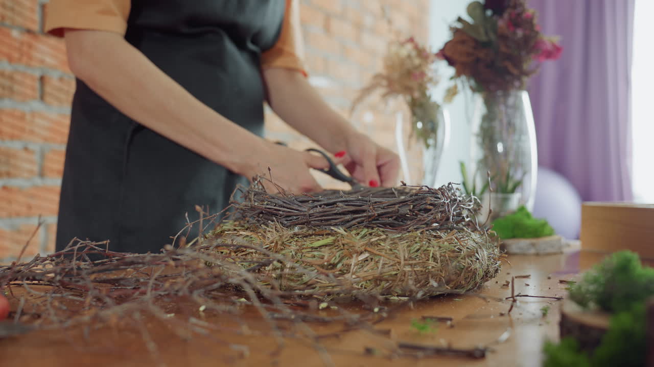 Female florist in apron arranging rustic wreath with twigs and hay on wooden table surrounded by dried flowers, moss and natural decorations, creating seasonal handmade craft emphasizing creativity