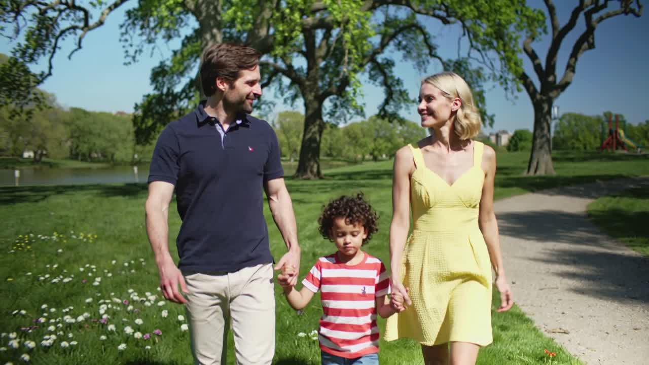 Genitori che tengono il bambino vicino, camminando verso l'obiettivo su un sentiero di ghiaia, vestiti in blu navy, giallo e a strisce