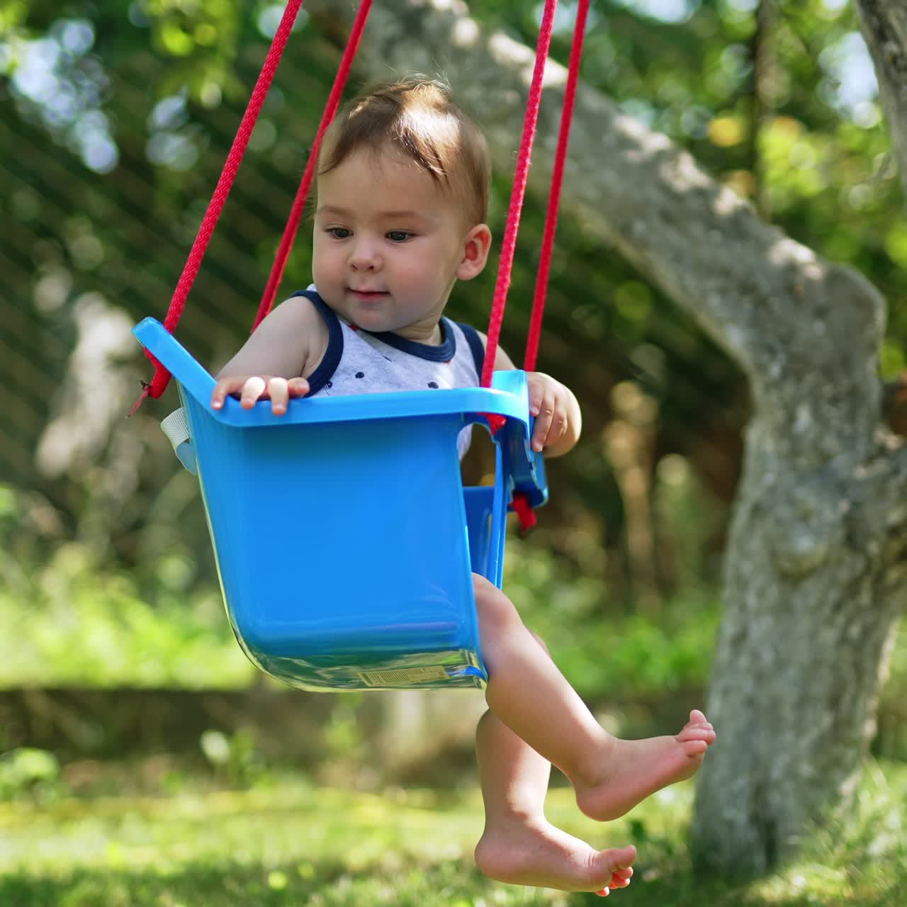 Parent's hand pushing the blue swing with cute baby sitting inside. Fun time with kid in the garden in summer. Nature backdrop
