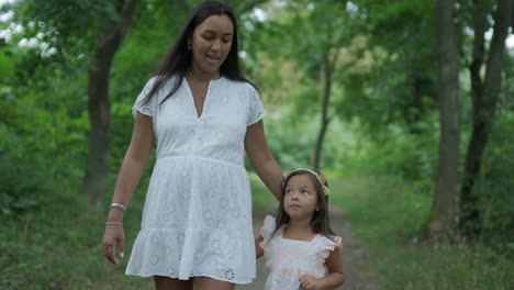 a pregnant woman and her young daughter, both dressed in white, walk hand-in-hand through a grassy park. the scene captures a serene moment of family bonding in nature, surrounded by trees.