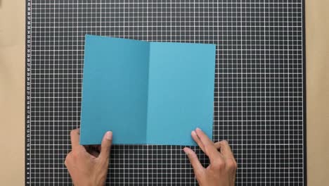 hands precisely folding blue cardboard on grid mat, crafting, attention to detail, top view