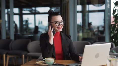 Una-Hermosa-Joven-Vestida-De-Negocios-Se-Sienta-En-Una-Oficina-Moderna,-Hablando-Por-Teléfono-Mientras-Trabaja-En-Su-Computadora-Portátil