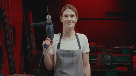 portrait of young woman in apron smiling at camera with turned on electrodrill in metal workshop